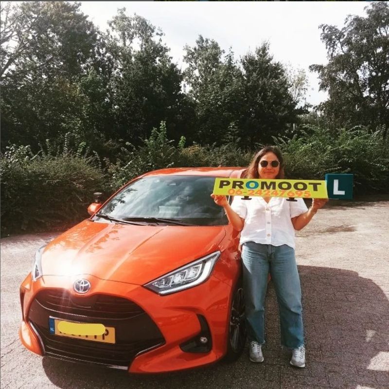 Geslaagde leerling met het PROMOOT-lesbord naast de oranje lesauto