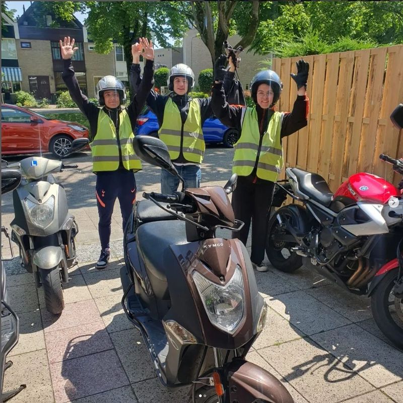 Drie leerlingen in veiligheidshesje met de hand omhoog naast de lesscooters van Autorijschool Promoot in Ridderkerk